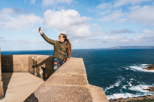 Молодая женщина, стоящая на берегу, делает селфи со смартфоном. На ней зеленая куртка и узорчатые леггинсы, с длинными волосами, дующими на ветру.. 