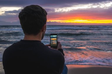 Deniz kenarında dururken canlı bir plaj günbatımını telefonuyla yakalayan bir adam.