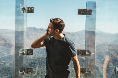 Manzaralı dağ vadisinin yukarısındaki cam platformda akıllı telefon kullanan genç adam.