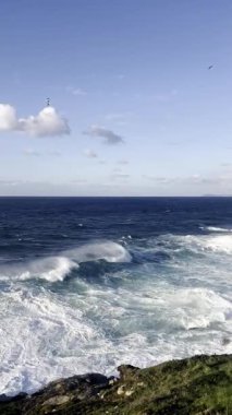 Dalgalar engebeli kayalıklara çarpıyor mavi gökyüzünün altında, güneşli bir sahil yolu ile bitiyor.