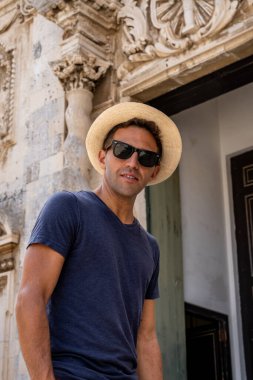 Sicilya Siracusa 'daki tarihi binanın önünde duran hasır şapkalı ve güneş gözlüklü bir adamın portresi.