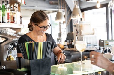 Modern bir barın arkasında çalışan genç bir kadın, bir şişe açarken gülümsüyor.