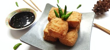 Fried milk tofu or Tahu susu goreng served on a plate and chopsticks with black sauce, green chili pepper on white background. Traditional snack from Indonesia clipart