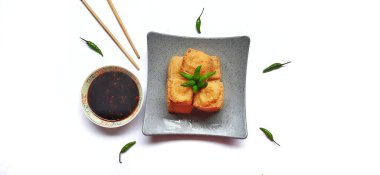 Fried milk tofu or Tahu susu goreng served on a plate and chopsticks with black sauce, green chili pepper on white background. Traditional snack from Indonesia clipart