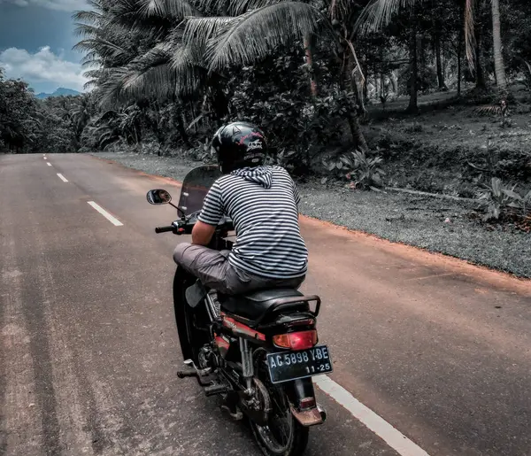 Yalnız bir motosikletçi yemyeşil ve yüksek palmiye ağaçlarıyla çevrili pürüzsüz, boş bir yolda ilerliyor. Açık yol ve sakin çevre macera ve özgürlük duygusunu çağrıştırıyor..