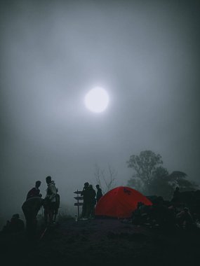 Sisli, atmosferik bir fotoğraf bir grup kampçıyı bir dağ yolunda sis kaplı bir gökyüzünün altında parlak kırmızı bir çadırın etrafında toplanırken gösteriyor. Güneş kalın sislerin arasından belli belirsiz parlıyor, engebeli ortama gizemli bir hava katıyor..