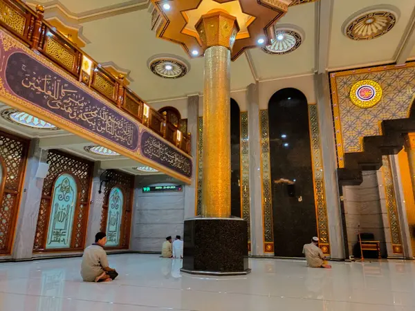 Büyük bir camide, bazı bireyler sessizce oturup dua ederler. İçerisi süslü bir şekilde İslami kaligrafi, altın sütunlar ve karmaşık mimari tasarımlarla süslenmiş. Huzurlu ortam sıcak ışıkla vurgulanır..