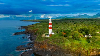 Okyanusa bakan kırmızı ve beyaz deniz feneri olan, mavi gökyüzünün altında yemyeşil bulutlarla çevrili manzaralı kıyı manzarası..