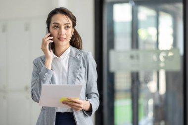 Genç gülümseyen Asyalı kadın cep telefonuyla konuşuyor, iş yerinde elinde belgelerle duruyor.