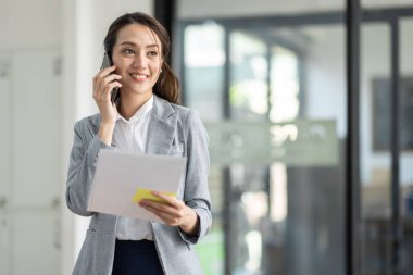 Genç gülümseyen Asyalı kadın cep telefonuyla konuşuyor, iş yerinde elinde belgelerle duruyor.