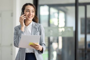 Genç gülümseyen Asyalı kadın cep telefonuyla konuşuyor, iş yerinde elinde belgelerle duruyor.