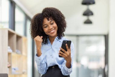 Başarılı ve heyecan verici genç Afro-Amerikan iş kadını telefon ekranına bakıyor, Afrikalı kadın iş yerinde akıllı telefonuyla kazanacağı için heyecanlı..