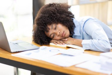 Exhausted millennial African woman sleeping on her office desk, next to laptop and documents, tired of overworking. Young African girl workaholic suffering from chronic fatigue at workplace clipart