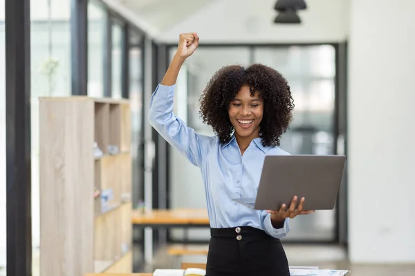 Excited happy Asian African woman looking at the laptop screen, celebrating an online win, overjoyed young African girl screaming with joy, isolated over a office background