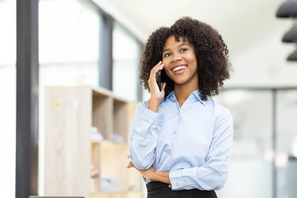 Bilgisayarlı dizüstü bilgisayar kullanan, telefonda konuşan ve iş yerinde dizüstü bilgisayarda çalışan mutlu Asyalı Afrikalı kadın, vergi konseptini belgeliyor.