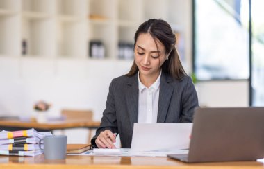 Neşeli Asyalı iş kadını çalışma alanı ofisinde çalışıyor. Ofisteki ahşap masada matematik finansmanı yapmak, vergi, muhasebe, finansal konsebe.