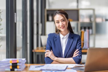 Kahverengi saçlı, açık mavi ceketli, gülümseyen Asyalı kadın ofiste evrak bilgisayarıyla çalışıyor, mali raporu analiz ediyor, iş planı yatırımı, finans analizi kavramını inceliyor..