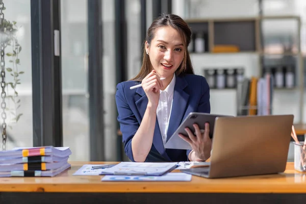 Kahverengi saçlı, açık mavi ceketli, gülümseyen Asyalı kadın ofiste evrak bilgisayarıyla çalışıyor, mali raporu analiz ediyor, iş planı yatırımı, finans analizi kavramını inceliyor..
