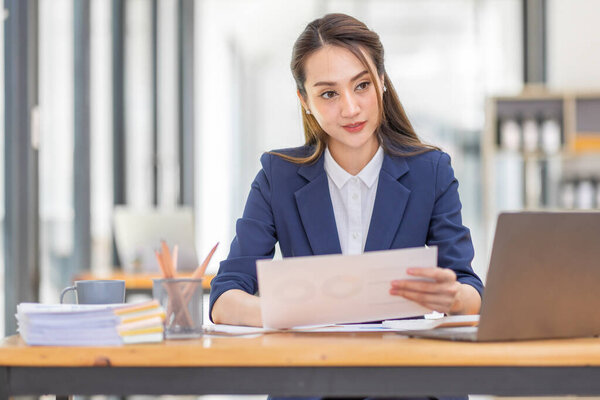 Brown haired wearing light blue jacket smiling Asian woman work with document laptop in office, doing planning analyzing the financial report, business plan investment, finance analysis concept.