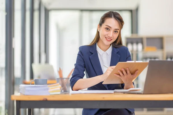 Kahverengi saçlı, açık mavi ceketli, gülümseyen Asyalı kadın ofiste evrak bilgisayarıyla çalışıyor, mali raporu analiz ediyor, iş planı yatırımı, finans analizi kavramını inceliyor..