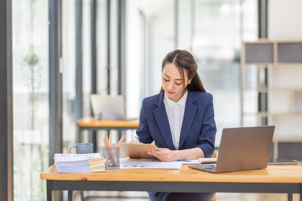 Brown haired wearing light blue jacket smiling Asian woman work with document laptop in office, doing planning analyzing the financial report, business plan investment, finance analysis concept.