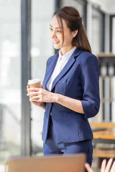 Portrait of young Asian woman hoding coffee cup and standing in a busy modern workplace