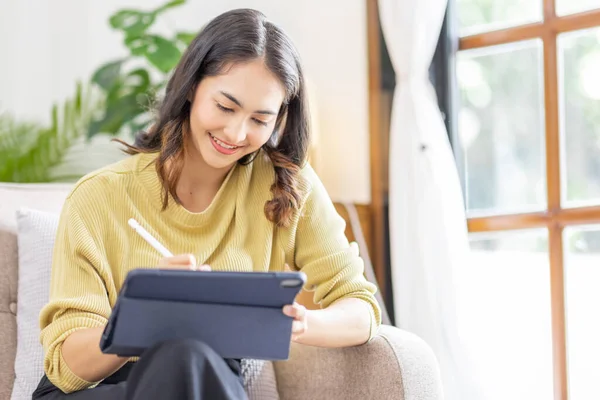 Remote learning concept. Happy Indian Asian Girl studying online, sitting on sofa with teblet at home. Indian teenager in Islamic headscarf using tablet, doing assignment, communicating to teacher,