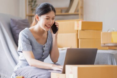 Gülümseyen Asyalı güzel kadın KOBİ girişimci işletme sahibi elinde karton kutular olan işyeri sahibi, ürünleri paketliyor ve evdeki dizüstü bilgisayarlarında müşterilerin siparişlerini kontrol ediyor, KOBİ teslimatı kavramı İnternet 'te.