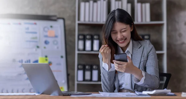 stock image Happy excited successful Asian businesswoman triumphing with smartphone and laptop at workplace.