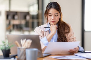 Image of young beautiful brooding Asian woman working with laptop while sitting at laptop in office, thinking of professional plan, project management, considering new business ideas.