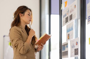 Young Business Asian woman using sticky notes and graph chart in glass wall analyzing strategy business plan to development grow to success