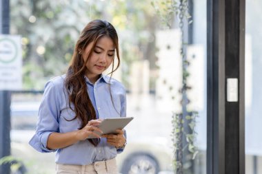 Young business Asian woman using tablet and standing near the window in workplace