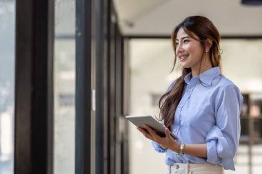Young business Asian woman using tablet and standing near the window in workplace