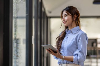 Young business Asian woman using tablet and standing near the window in workplace