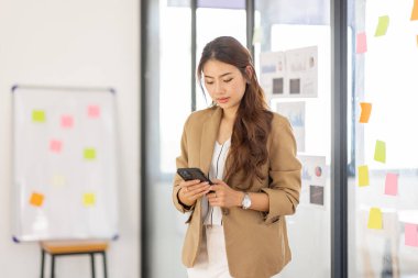 Ofiste telefonu kullanan gülümseyen Asyalı kadın. Cep telefonuna bakıp gülümseyen küçük bir girişimci..