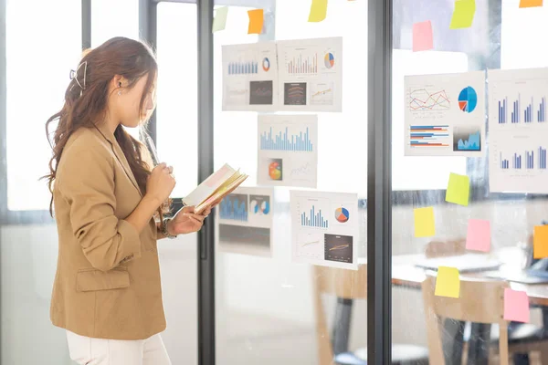 Young Business Asian woman using sticky notes and graph chart in glass wall analyzing strategy business plan to development grow to success