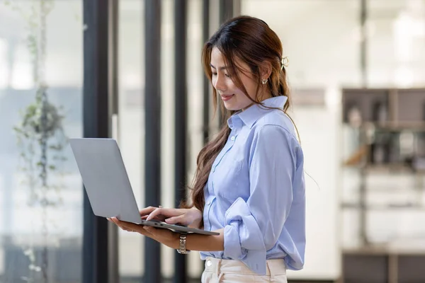 Portrait of Young business Asian woman using laptop computer, standing near the window in workplace