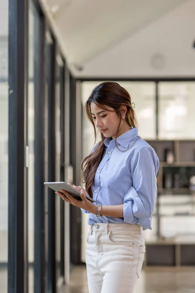 Young business Asian woman using tablet and standing near the window in workplace