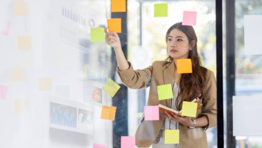 Young Business Asian woman using sticky notes and graph chart in glass wall analyzing strategy business plan to development grow to success