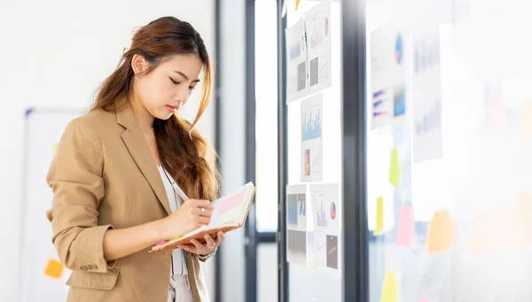 Young Business Asian woman using sticky notes and graph chart in glass wall analyzing strategy business plan to development grow to success