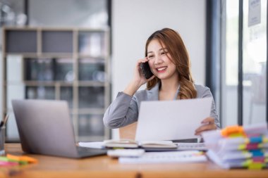 Smiling young Asian woman sitting and call phone device, looking at laptop, checking modern apps, texting messages, browsing internet doing shopping relaxing at home.