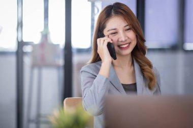 Smiling young Asian woman sitting and call phone device, looking at laptop, checking modern apps, texting messages, browsing internet doing shopping relaxing at home.