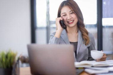 Smiling young Asian woman sitting and call phone device, looking at laptop, checking modern apps, texting messages, browsing internet doing shopping relaxing at home.