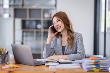 Smiling young Asian woman sitting and call phone device, looking at laptop, checking modern apps, texting messages, browsing internet doing shopping relaxing at home.