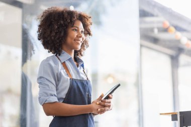 Successful african woman in apron standing coffee shop door. Happy small business owner holding tablet and working. Smiling portrait of SME entrepreneur seller business standing with copy space.