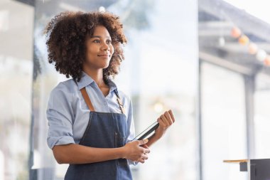 Successful african woman in apron standing coffee shop door. Happy small business owner holding tablet and working. Smiling portrait of SME entrepreneur seller business standing with copy space.