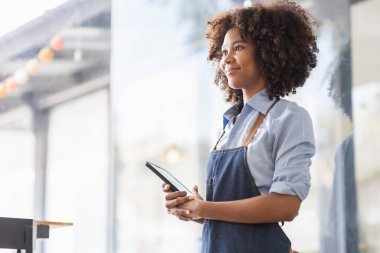 Successful african woman in apron standing coffee shop door. Happy small business owner holding tablet and working. Smiling portrait of SME entrepreneur seller business standing with copy space.