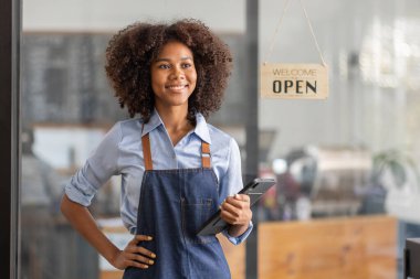 Successful african woman in apron standing coffee shop door. Happy small business owner holding tablet and working. Smiling portrait of SME entrepreneur seller business standing with copy space.