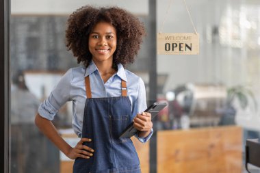 Successful african woman in apron standing coffee shop door. Happy small business owner holding tablet and working. Smiling portrait of SME entrepreneur seller business standing with copy space.