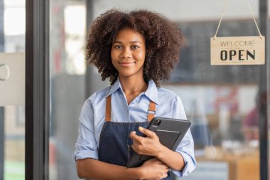 Successful african woman in apron standing coffee shop door. Happy small business owner holding tablet and working. Smiling portrait of SME entrepreneur seller business standing with copy space.